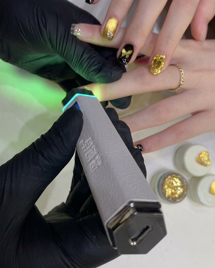 A gloved hand holds a portable handheld UV/LED lamp over a fingernail painted with black gel polish.
