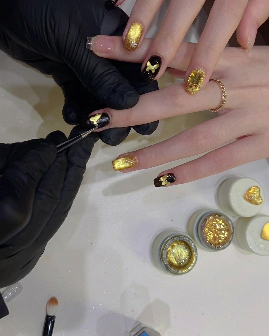 Precision tweezers peeling a butterfly-shaped stencil off a nail, revealing a glittering gold butterfly design underneath a black background.