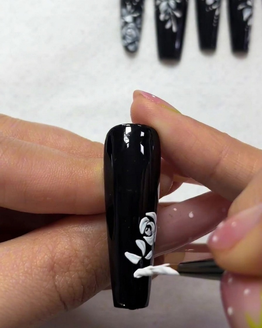 A close-up showing a fine brush painting small, tapered white leaf shapes near the base of a rose nail art design.