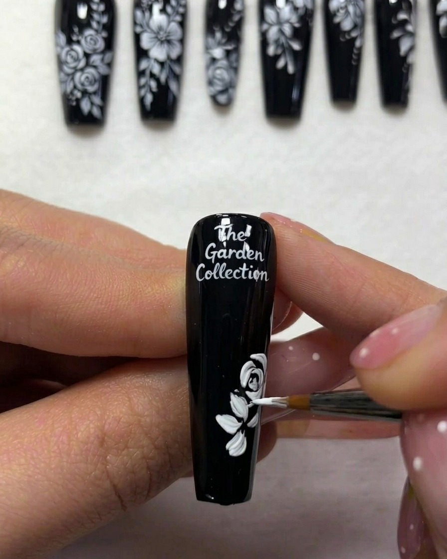 A close-up view of a nail artist adding thin white lines to represent stems extending from foliage on a black-painted nail.