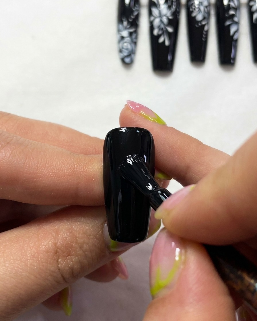 A close-up of a brush painting the thin, elegant white outlines of a butterfly wing structure on a black nail.
