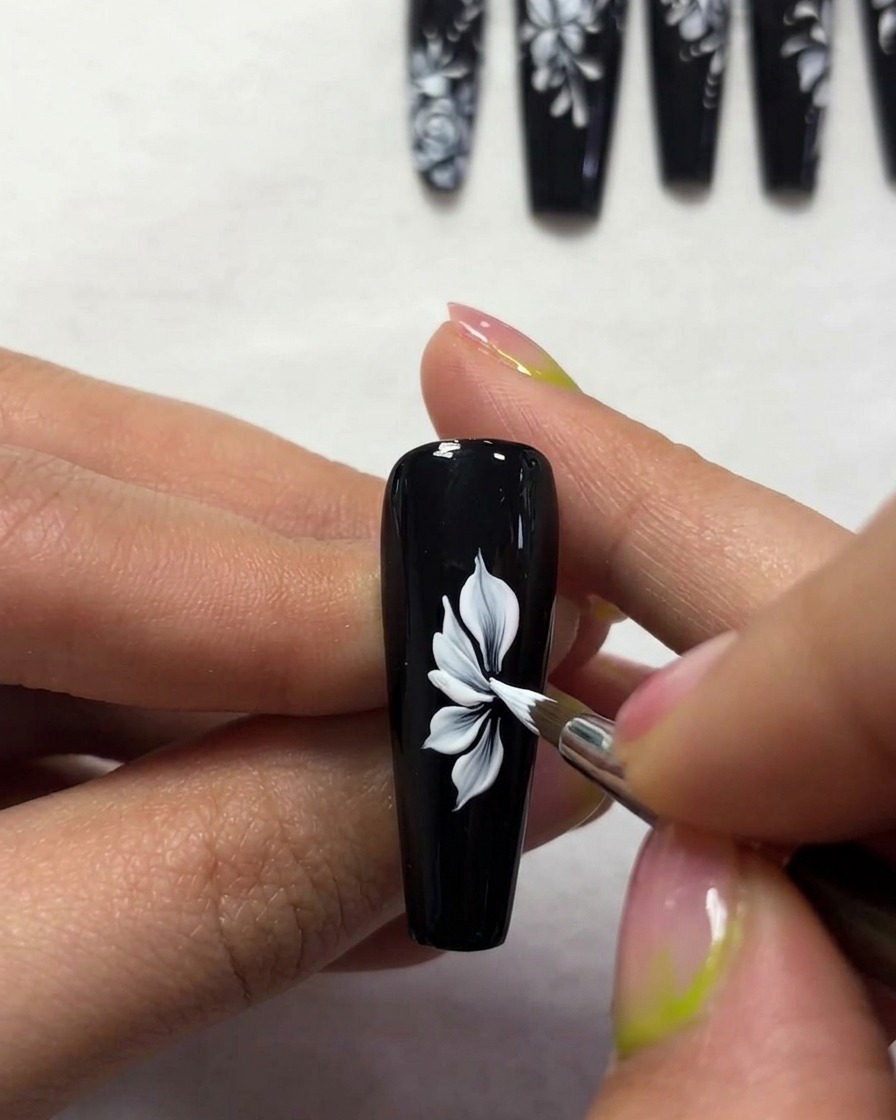 A close-up of a brush creating thin, elegant white butterfly wing outlines on a black nail surface.