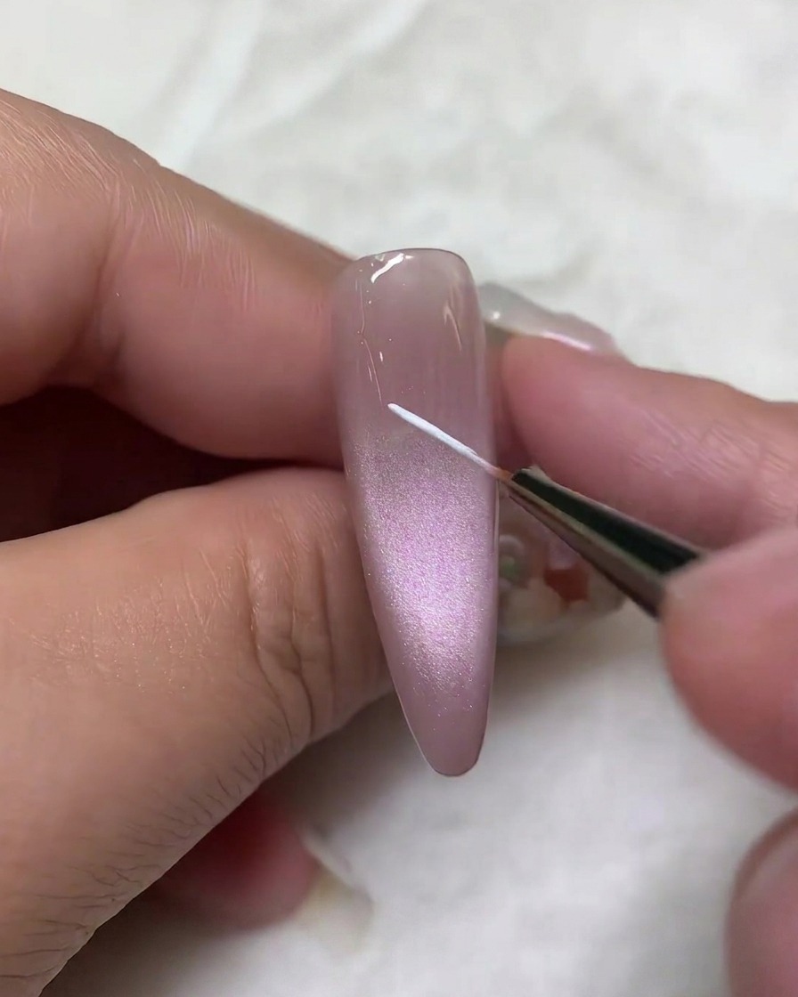 A close-up view of a fine liner brush applying a thin white line vertically along the center of a mauve cat-eye gel nail.