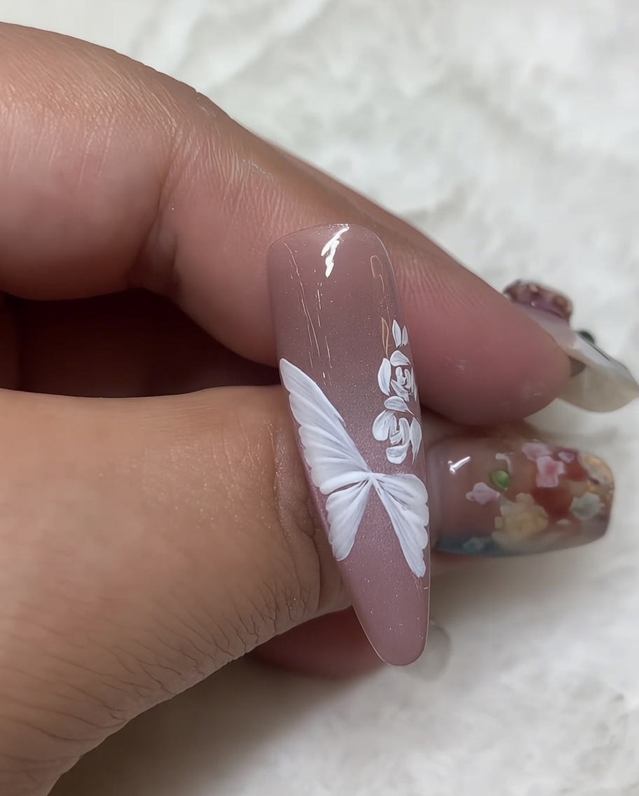 A close-up of a nail with a white gel-painted rose and a fine liner brush adding small, detailed white leaves to the side of the floral design.