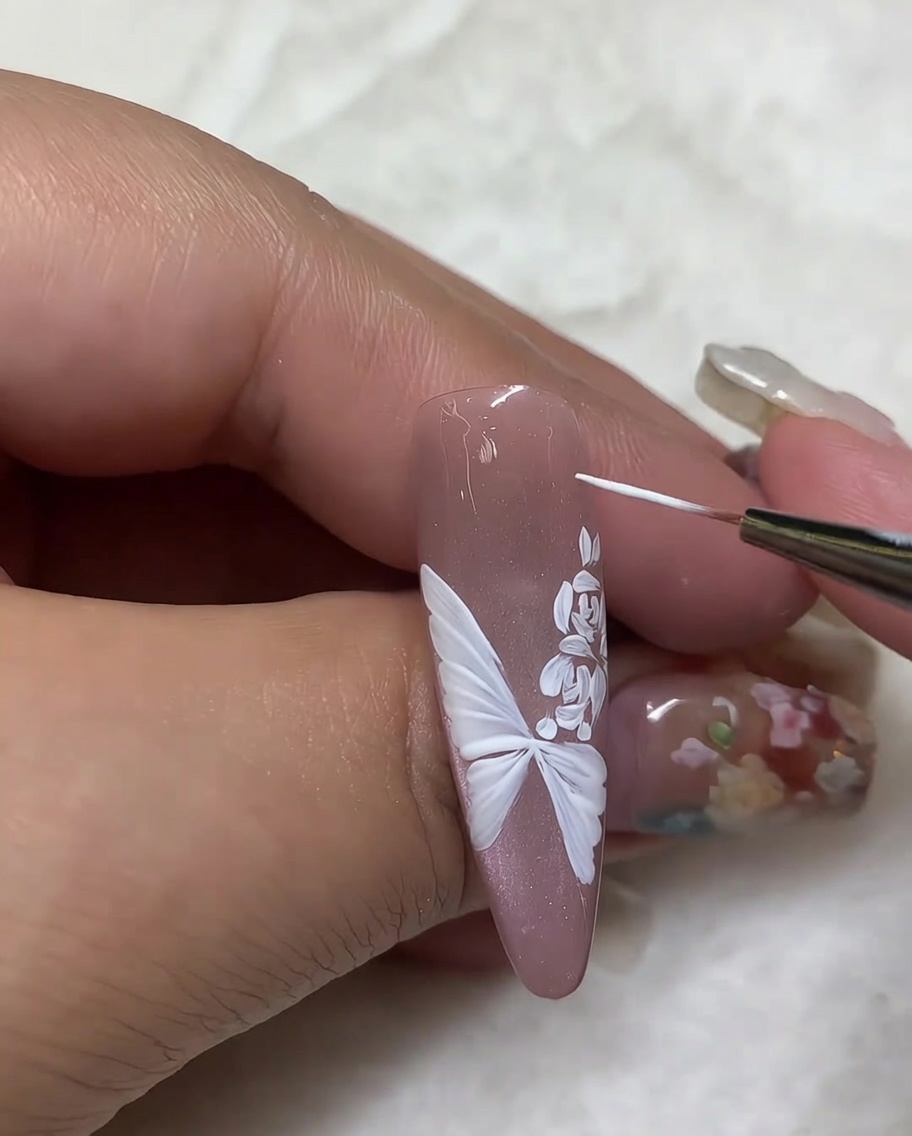 A close-up view showing a fine liner brush adding small white highlight dots around a butterfly and rose design on a nude-colored nail.