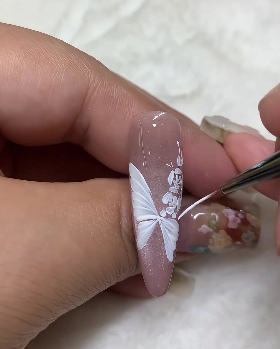 A close-up of a nail showing a fine liner brush tracing the outer edges of a white butterfly wing design to refine the detail.