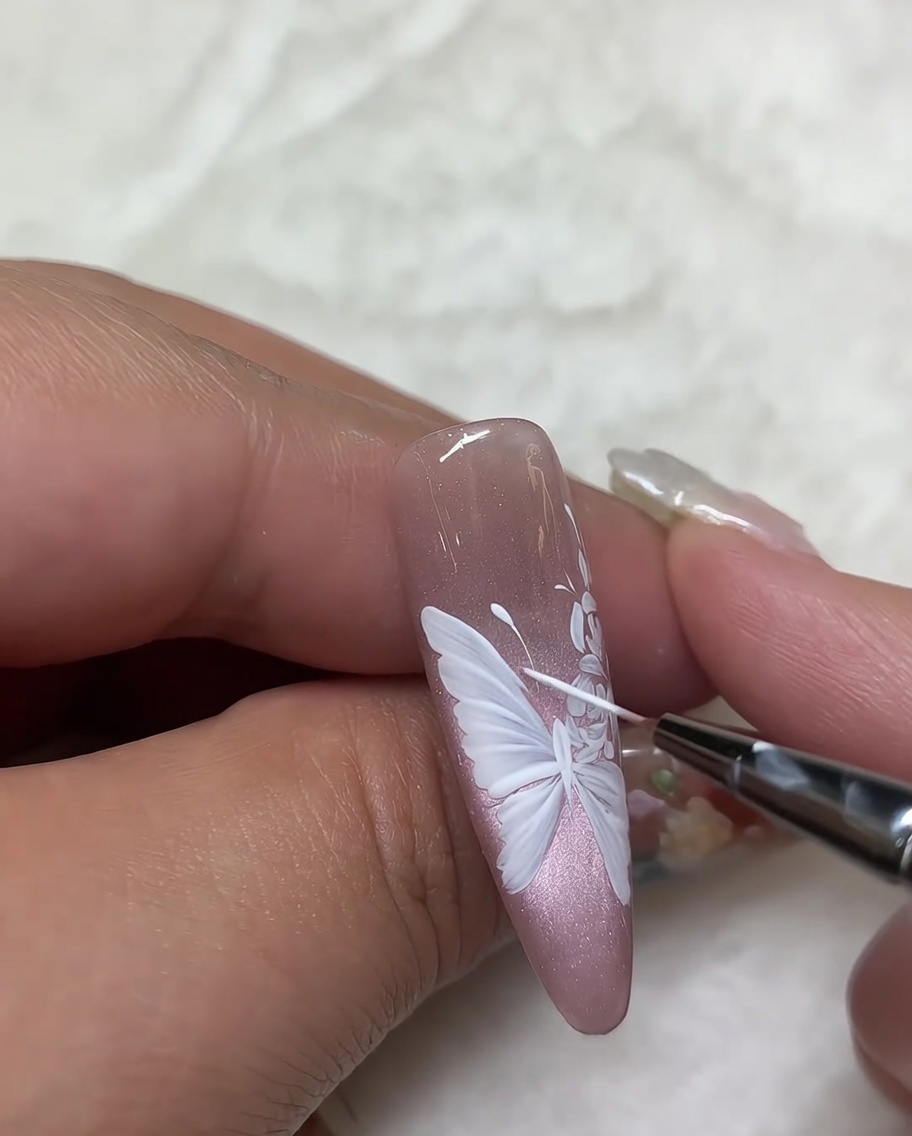 A close-up shot of a nail featuring a white rose design, with a fine liner brush being used to perform final touch-ups on the petal details.