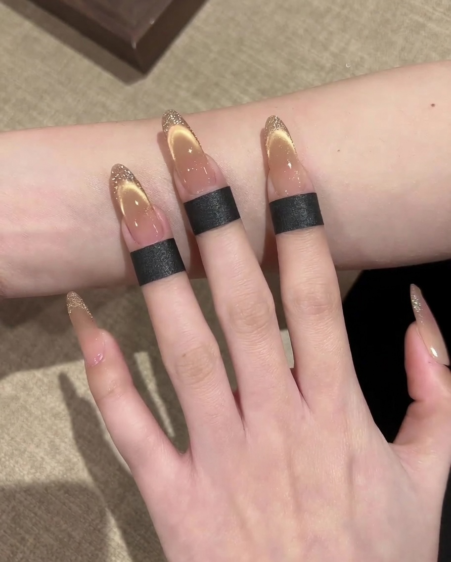 A close-up of a hand using a thin liner brush to paint a gold glitter outline along the edge of a French-style cat-eye nail.