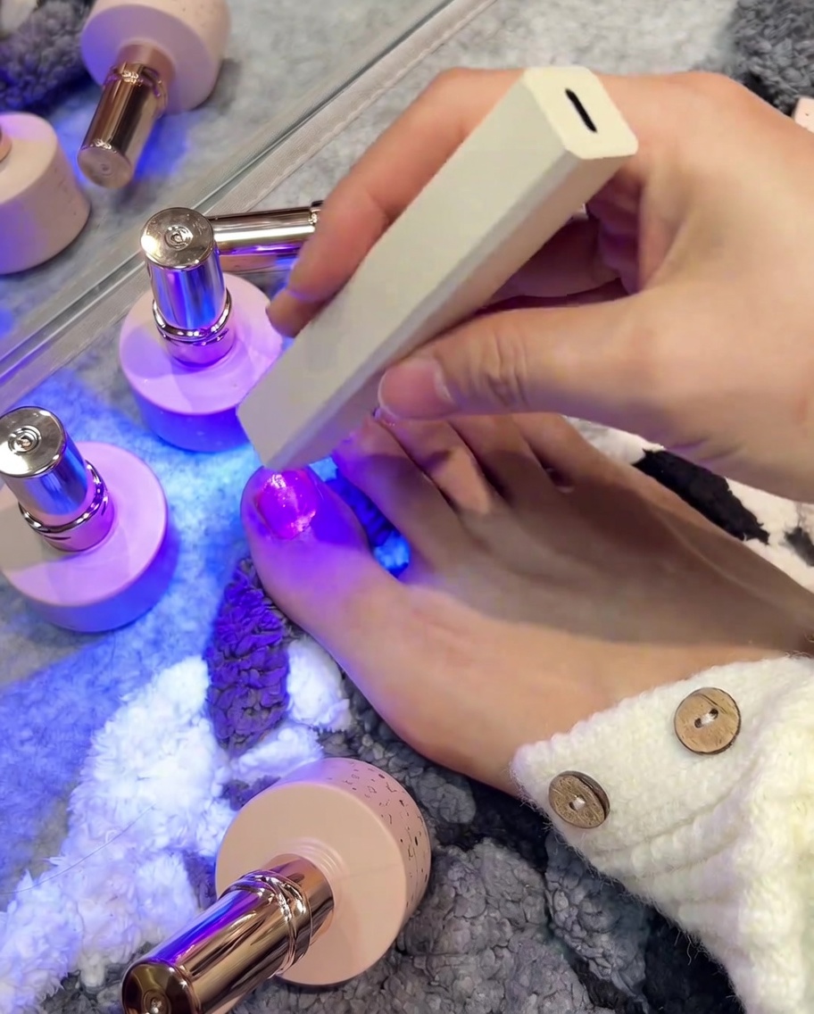 A hand holding a small white, rectangular UV/LED lamp directly over a freshly polished right big toenail to cure the gel.
