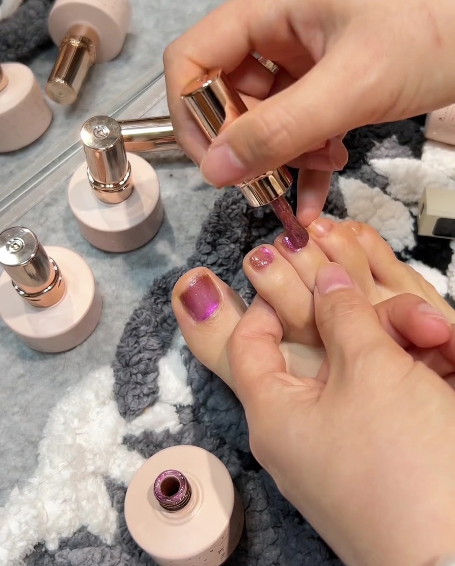 A brush applying rose pink gel polish to a toenail, followed by a magnet being used to create a cat-eye effect on the wet surface.