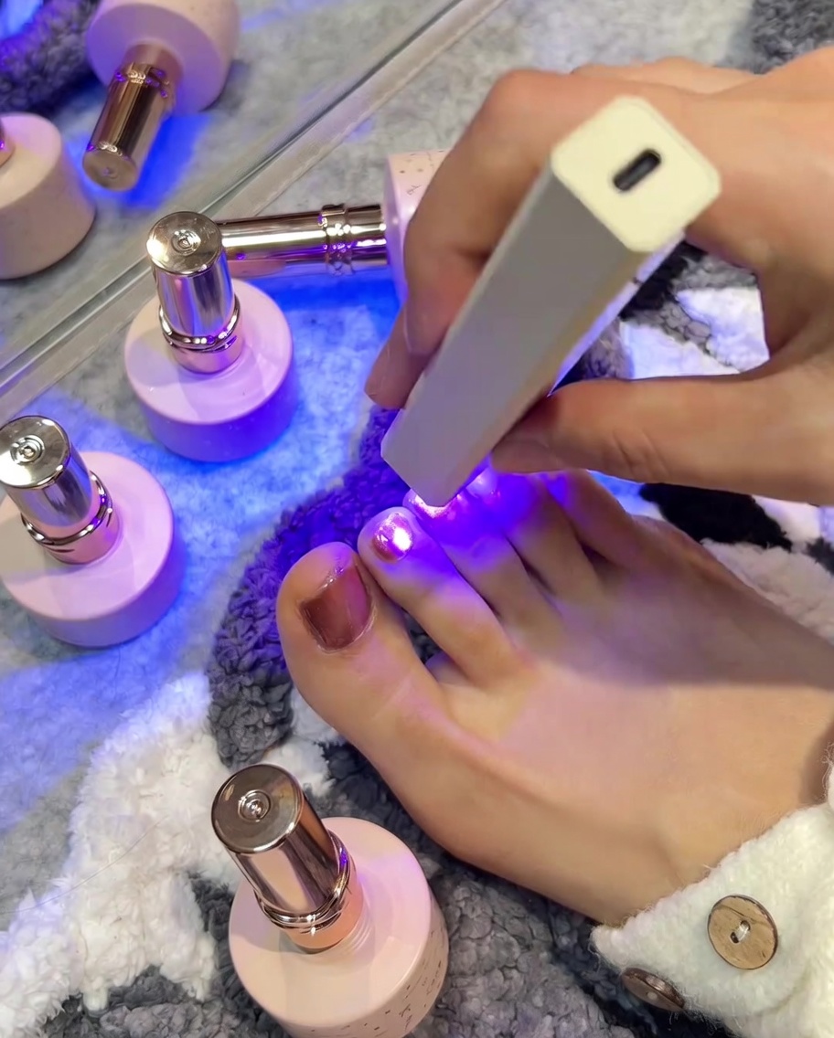 A handheld UV/LED lamp positioned over a pink toenail, with the violet light illuminating the nail as it cures.