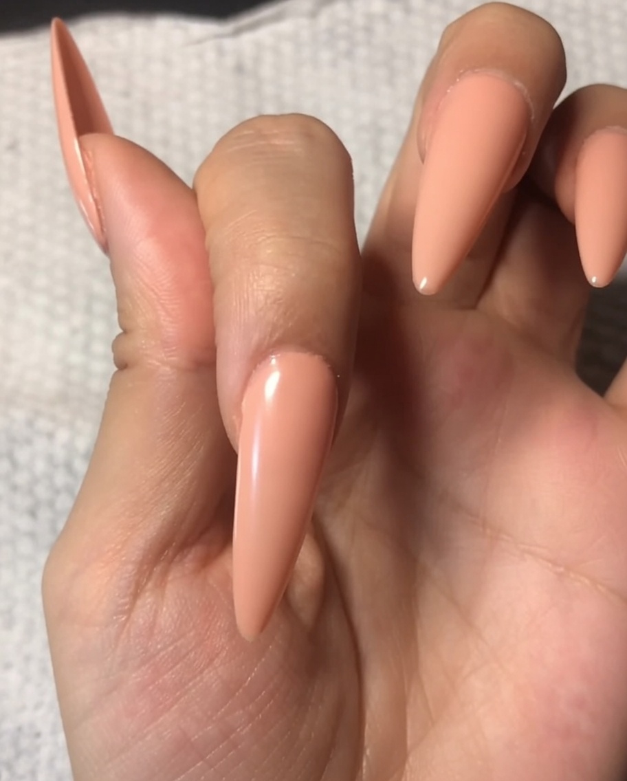 A brush applying a clear, glossy top coat to a fingernail already painted with a soft nude base.