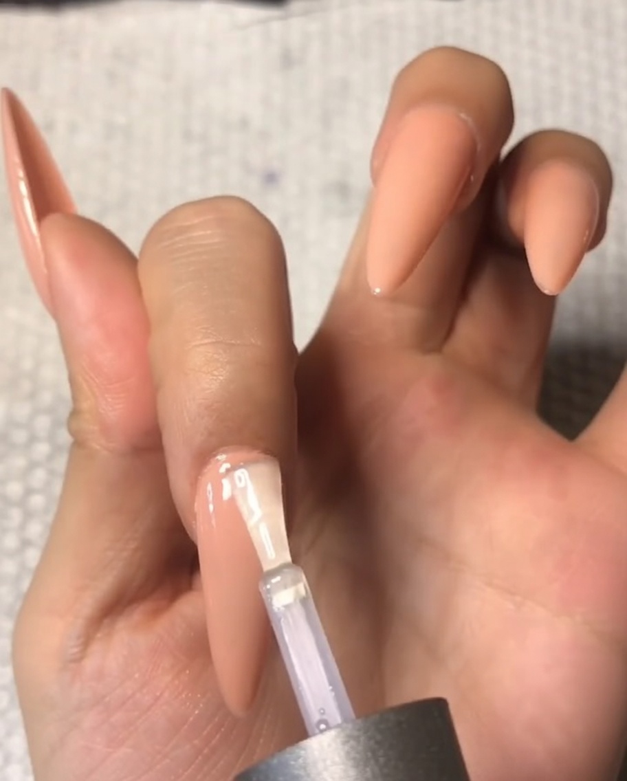 A close-up of a brush carefully applying a clear tempered top coat near the cuticle area of a nude almond-shaped middle fingernail.