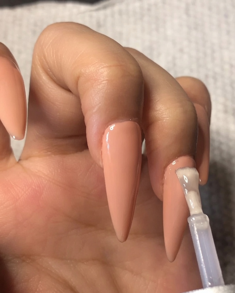 A brush applying a layer of clear gel top coat over a nude gel polish on the ring fingernail.
