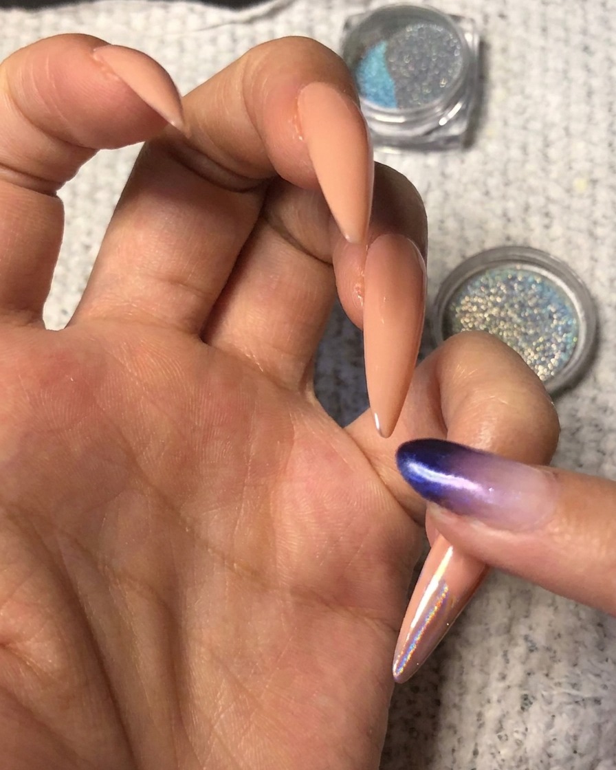 A finger pressing vibrant blue-purple holographic chrome powder onto the tip of a nude almond-shaped fingernail.
