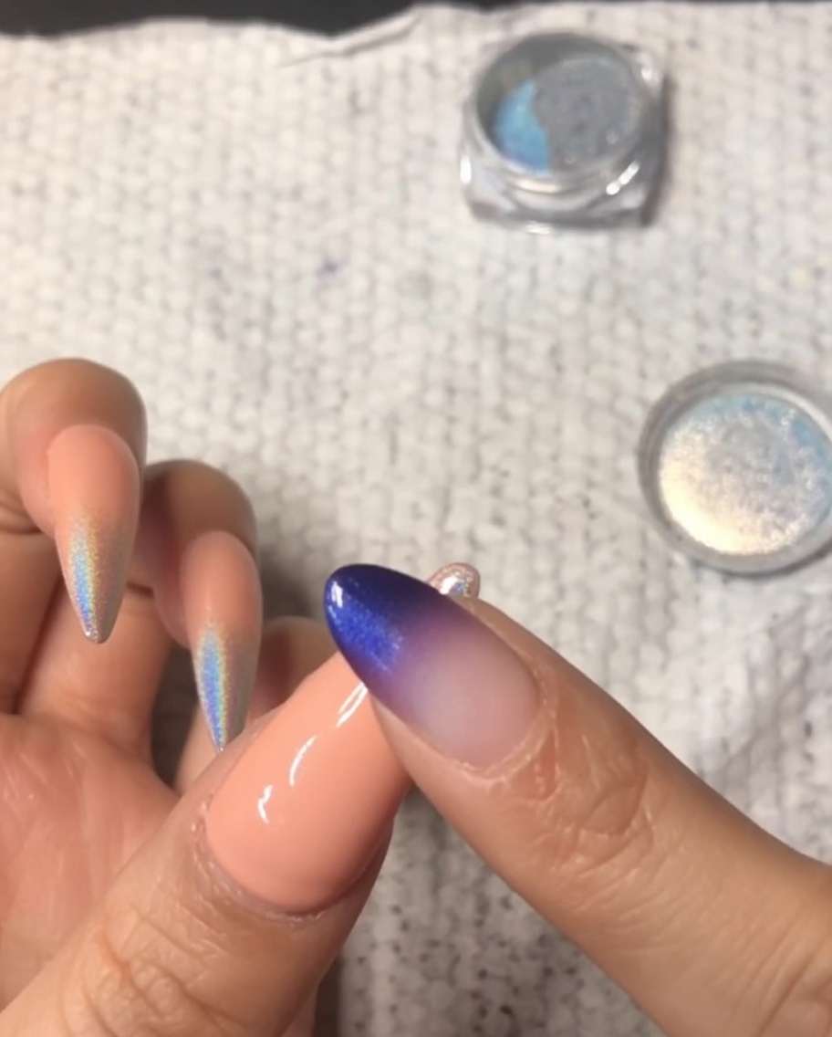 A close-up of a finger rubbing holographic chrome powder onto the tip of a pinky nail, creating an ombre gradient.