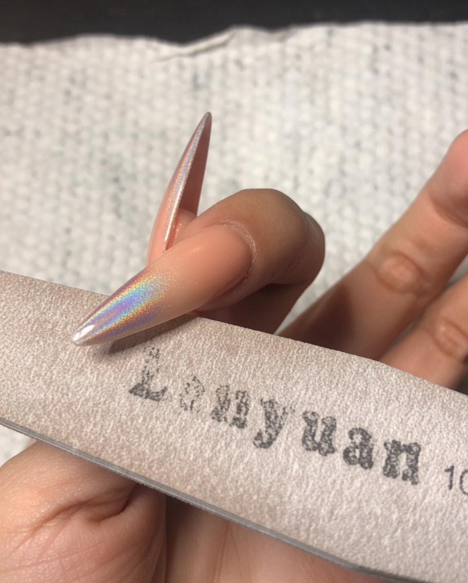 A hand using a fine sponge nail file to gently buff the free edge of a stiletto nail decorated with holographic chrome powder.