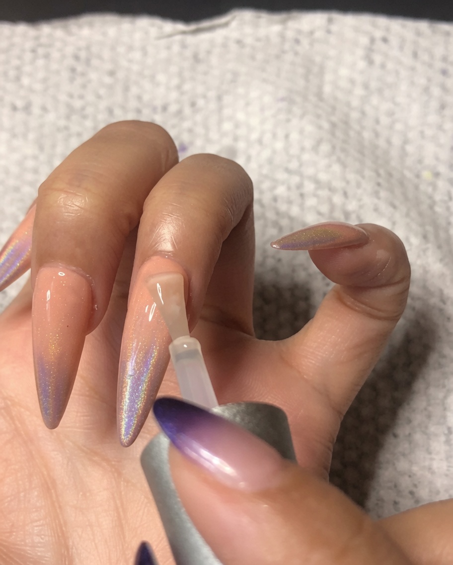 A nail brush applying a high-gloss top coat to the ring finger, sealing the holographic ombre nail art.