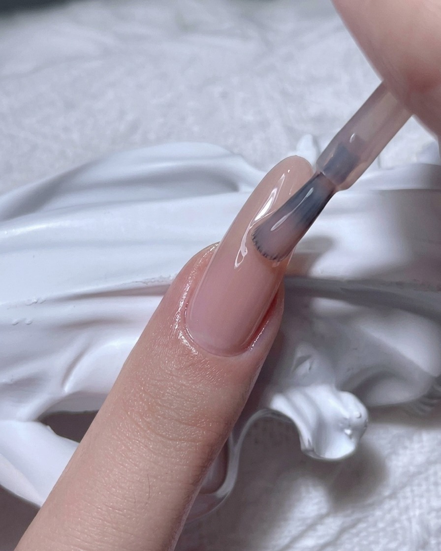 A brush applying a sheer nude gel polish onto a natural fingernail.