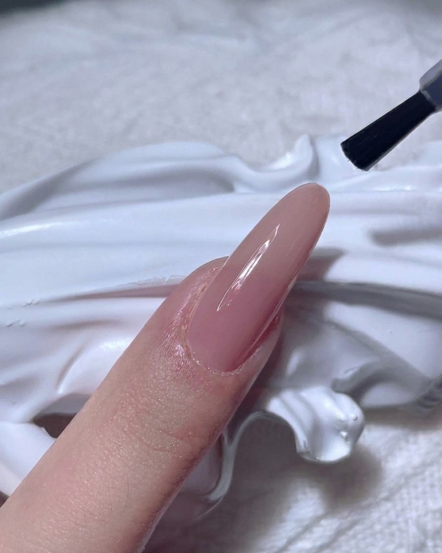 A nail brush applying a clear, shiny top coat over a nude-colored nail.