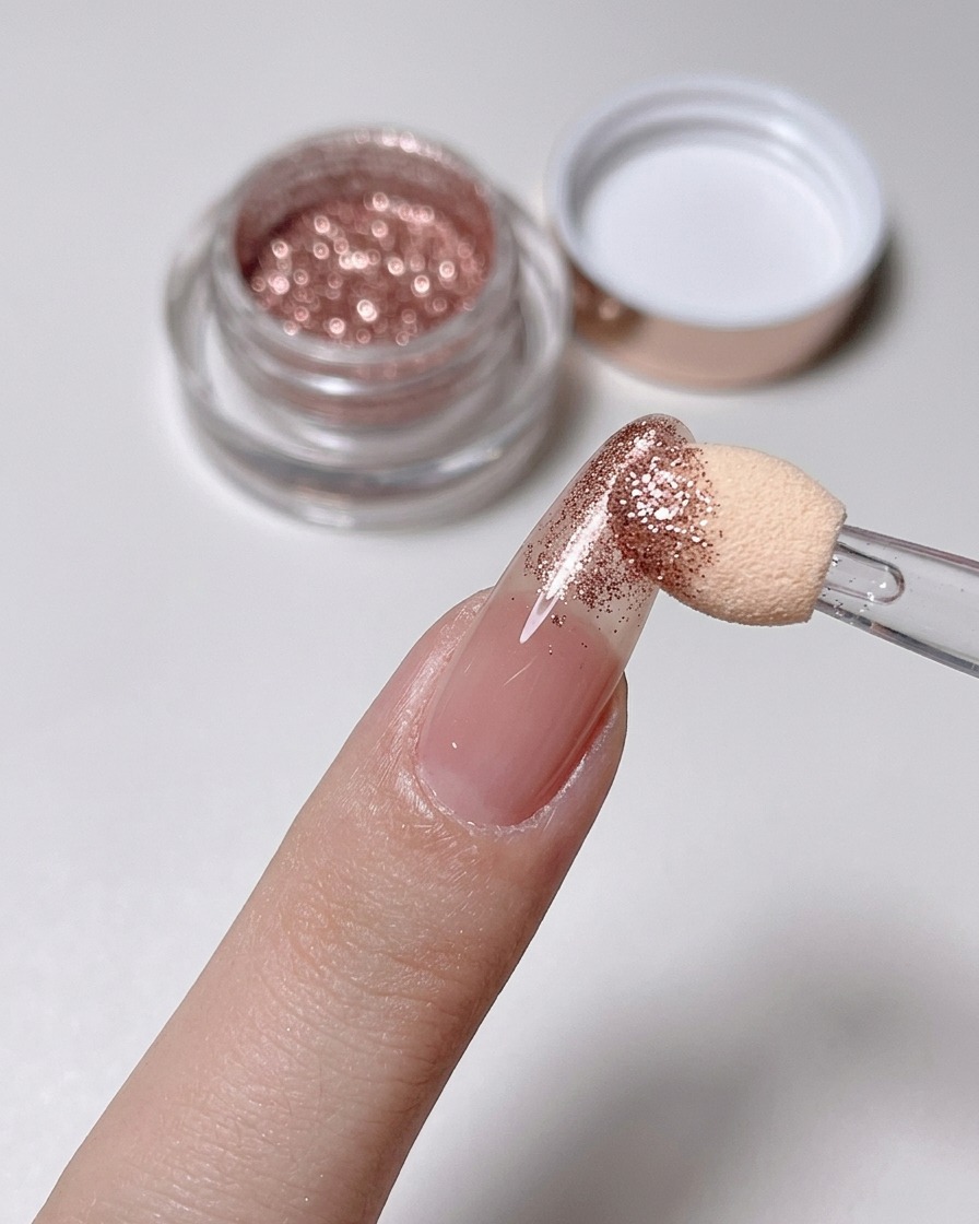 A sponge applicator buffing rose gold powder onto the tip of a nude nail.