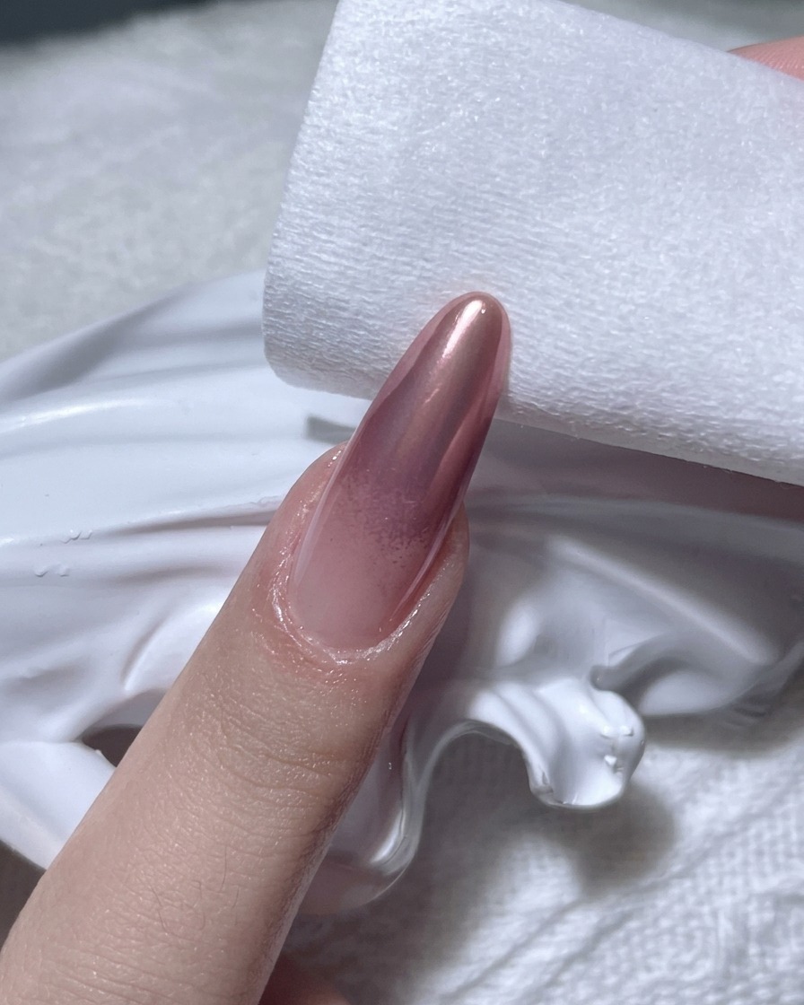A close-up shot of a hand using a lint-free pad to wipe the edges of a rose gold chrome-finished nail.