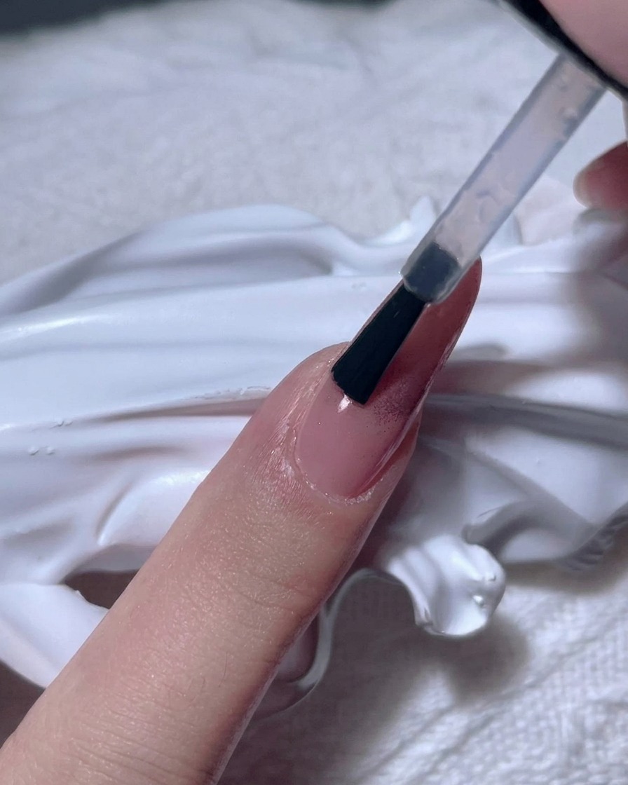 A brush applying a thick, clear gel over a rose gold chrome nail.
