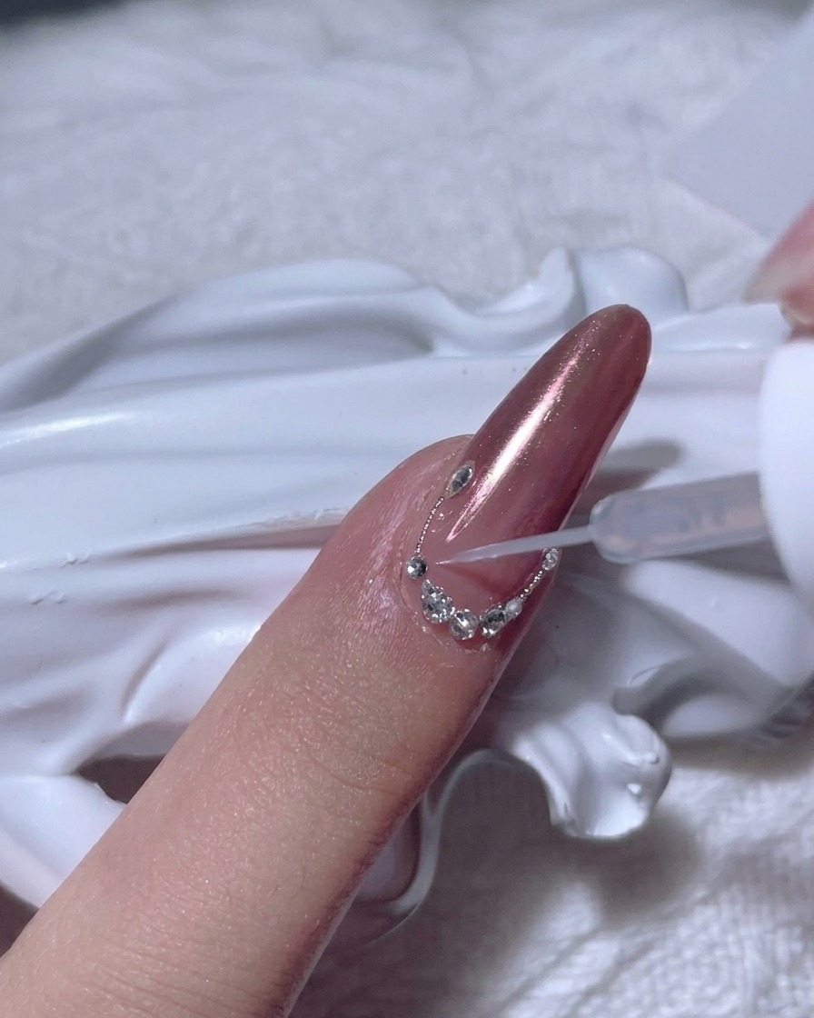 A completed arc of small clear rhinestones arranged along the cuticle line of a rose gold chrome nail.