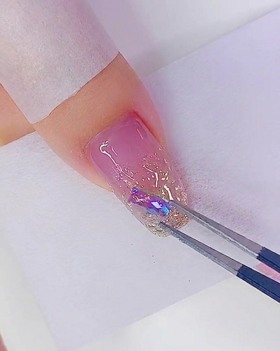 Tweezers placing an iridescent rhinestone onto a dot of adhesive on a glittery nail.