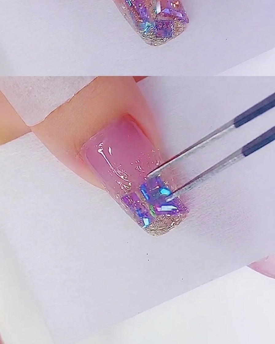 A close-up shot of tweezers placing iridescent rhinestones onto a nail tip that is already covered in rose gold glitter gel.
