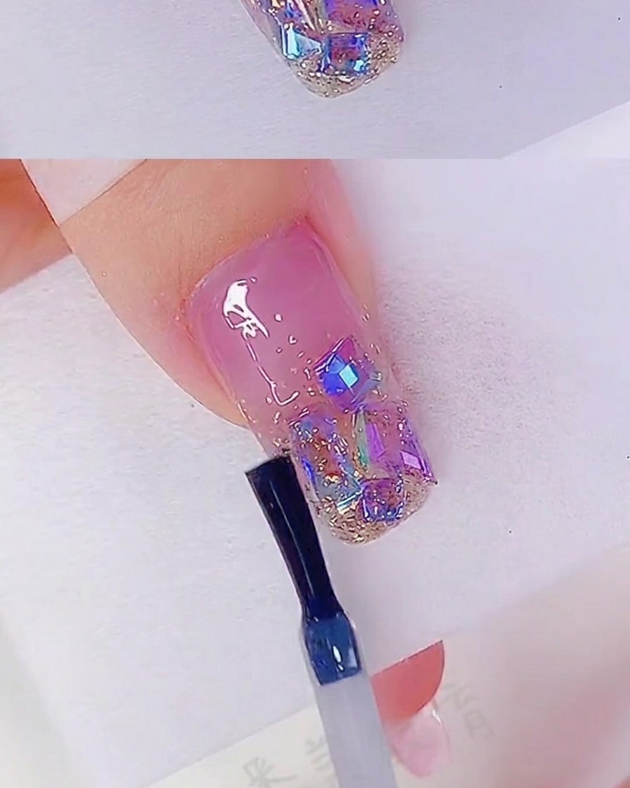 A nail brush applying a clear top coat over a nail bed decorated with rose gold glitter and iridescent rhinestones.