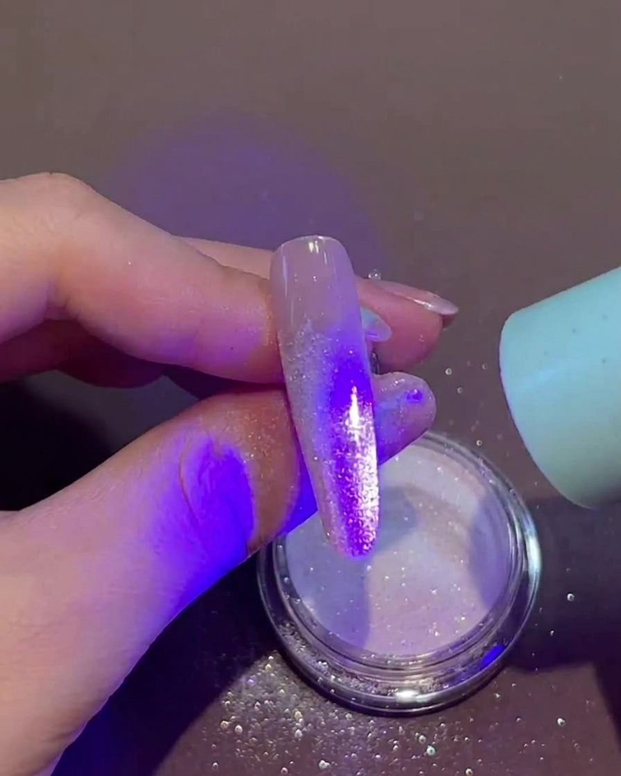 A hand holding a small portable UV/LED lamp directly over a fingernail that has wet gel and loose powder on the tip.