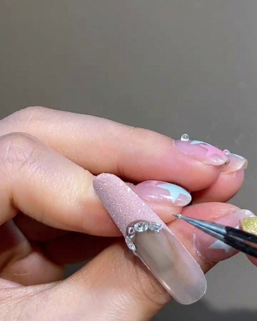 A close-up of tweezers placing a small silver metal bead along the boundary of a pink French tip on a manicured nail.