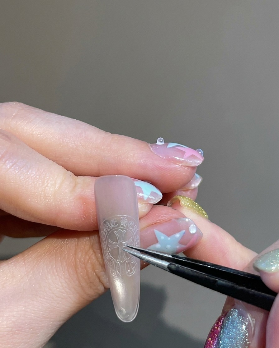 Tweezers holding and placing a cross-shaped sticker onto the center of a fingernail.