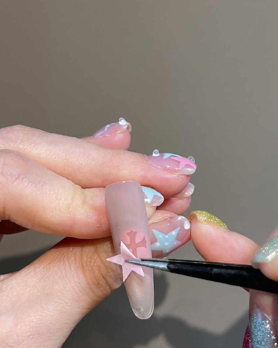 Tweezers peeling a sticker off a nail, leaving a clear cross-shaped silhouette in the center of the blush-toned nail.