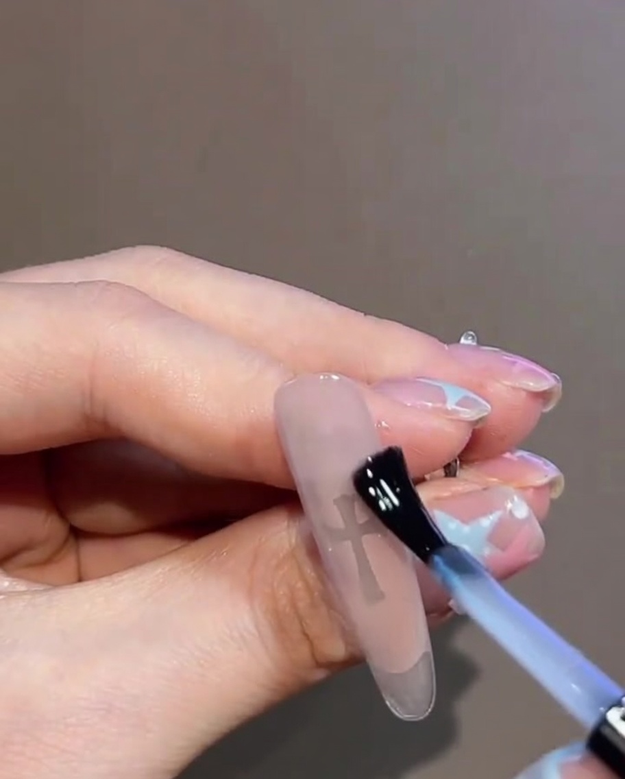 A close-up of a clear gel top coat being applied over a finished nail design featuring a cross sticker and rhinestones.