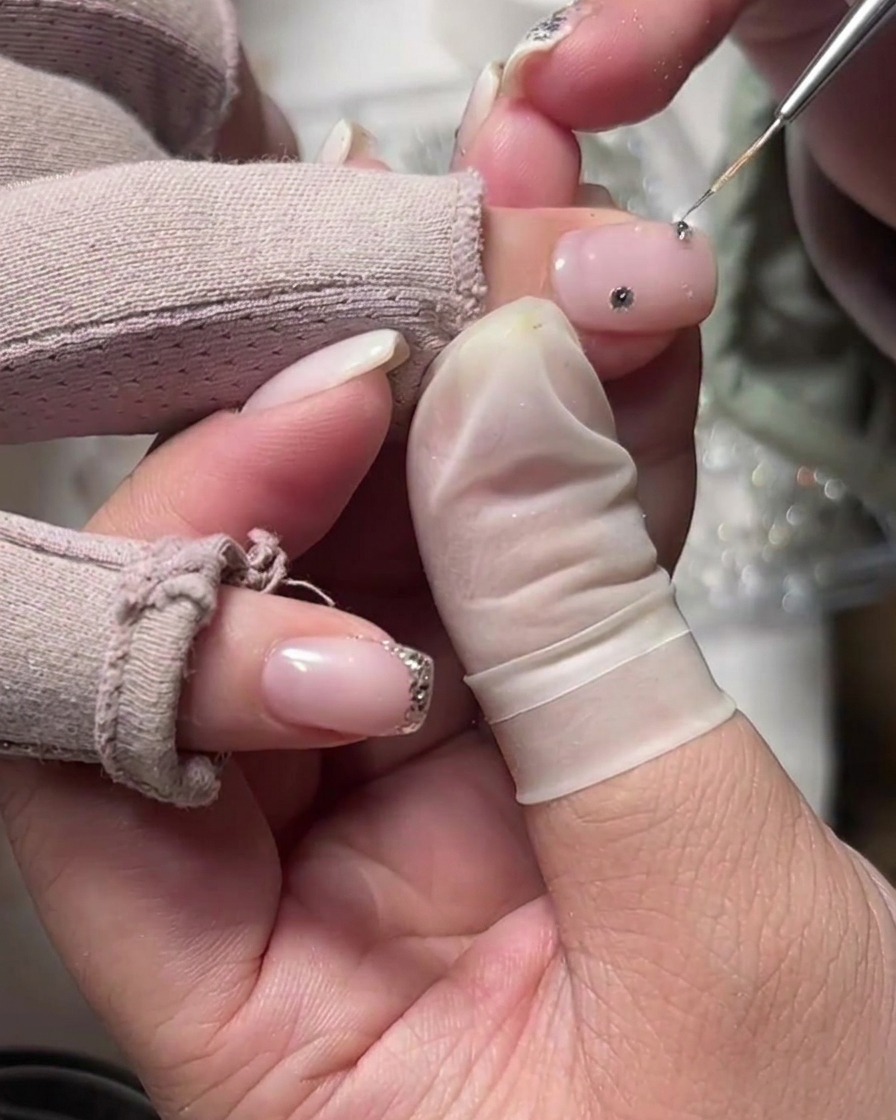 A fine-tipped tool placing a single rhinestone onto a milky white base nail near the cuticle area.