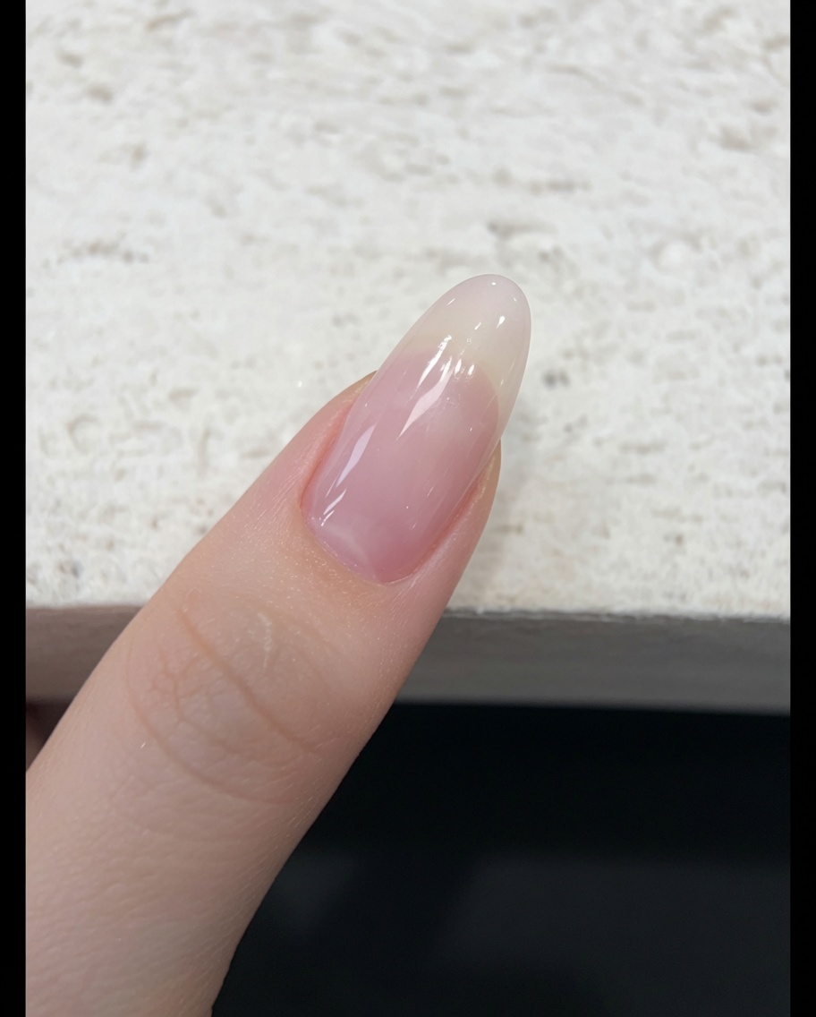 A brush applying a sheer, nude-pink jelly gel polish to a natural fingernail.