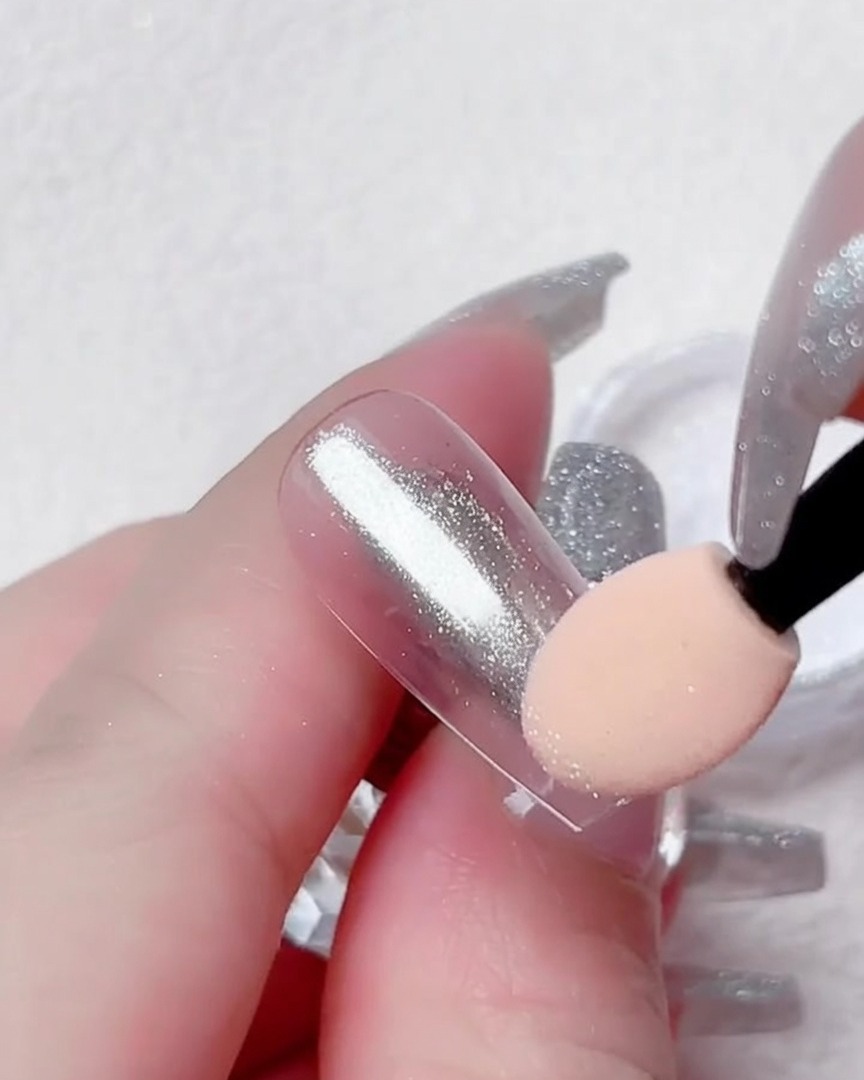 A close-up of a finger holding a clear nail tip while an eyeshadow sponge applicator brushes silver chrome powder onto its surface.