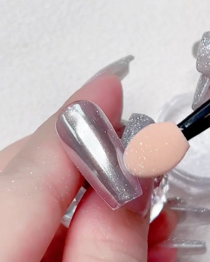 A close-up view of an applicator buffing silver chrome powder onto the edges of a clear nail tip.