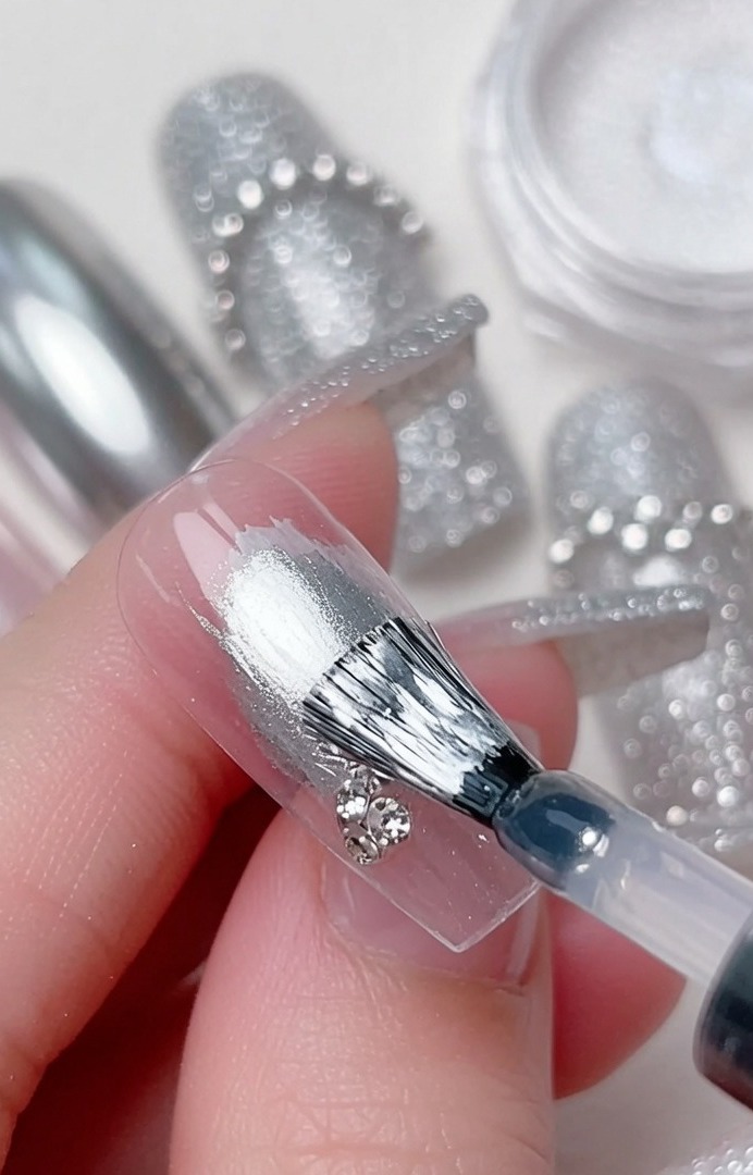 A detail brush placing a small, clear sparkling rhinestone at the intersection of clear gel swirls on a nail.