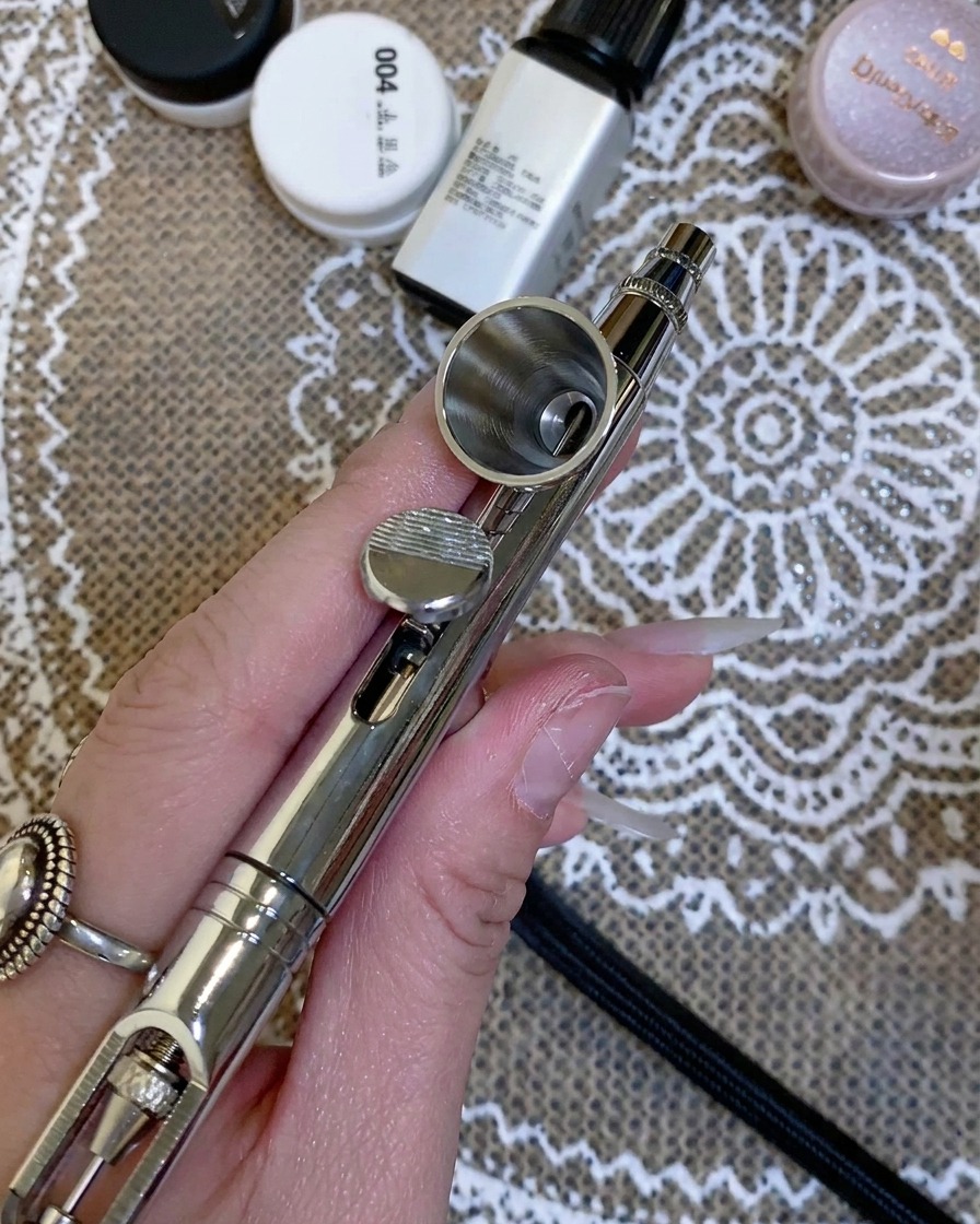 A close-up of a hand holding an airbrush, with clear liquid being dropped into the metal reservoir.