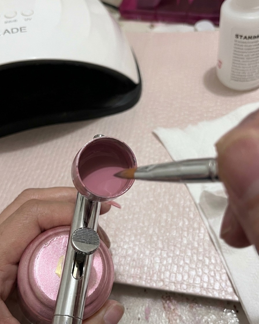 A close-up view of a metal mixing tool stirring pink gel polish inside an airbrush cup.
