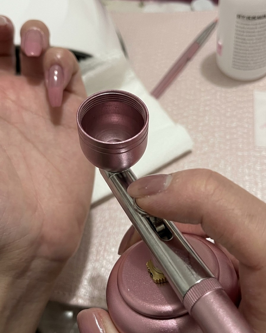 An airbrush applying a light dusting of peach-pink gel to the center of a prepped nail.