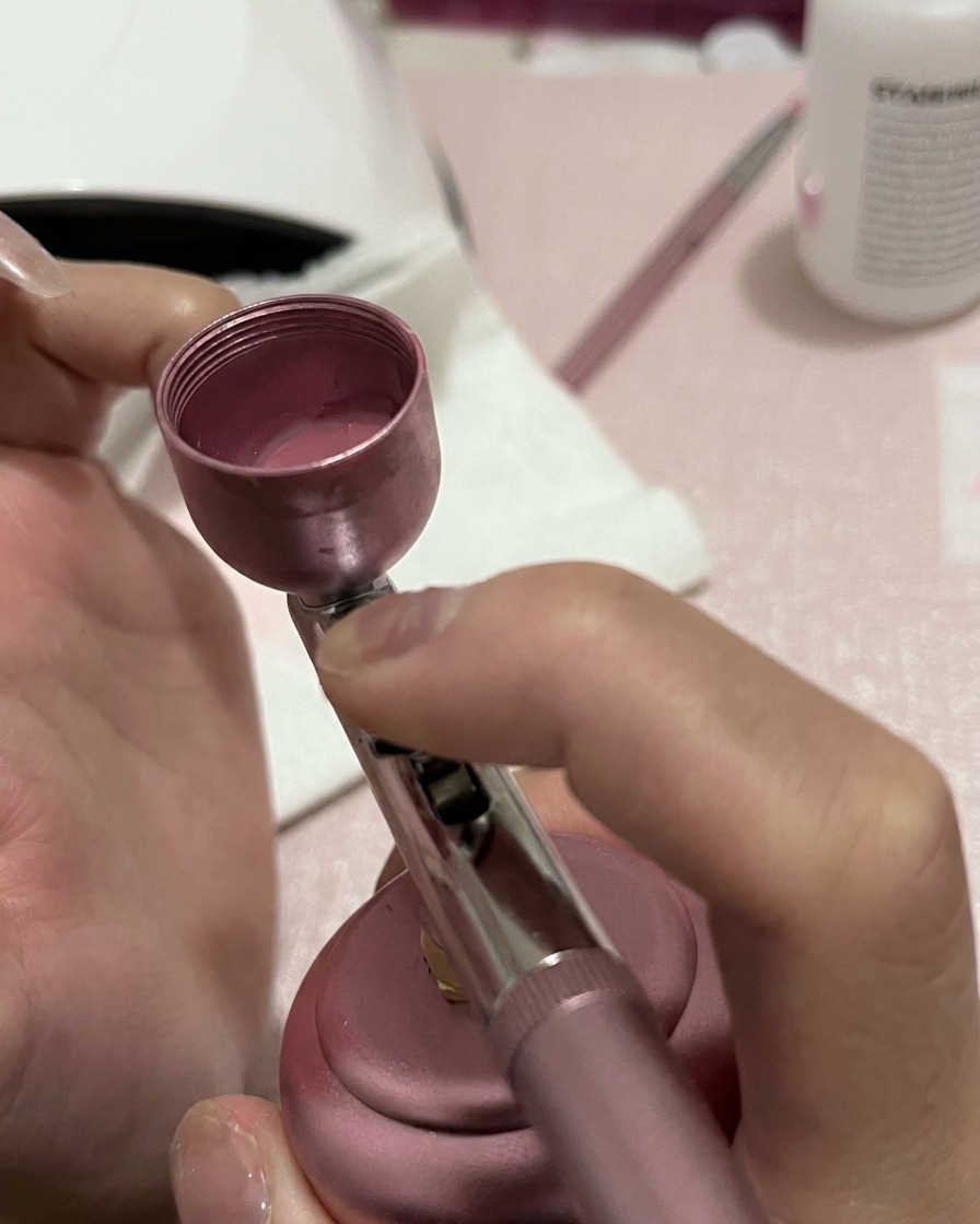 A close-up of a nail receiving a more concentrated spray of pink pigment at the base and mid-section.