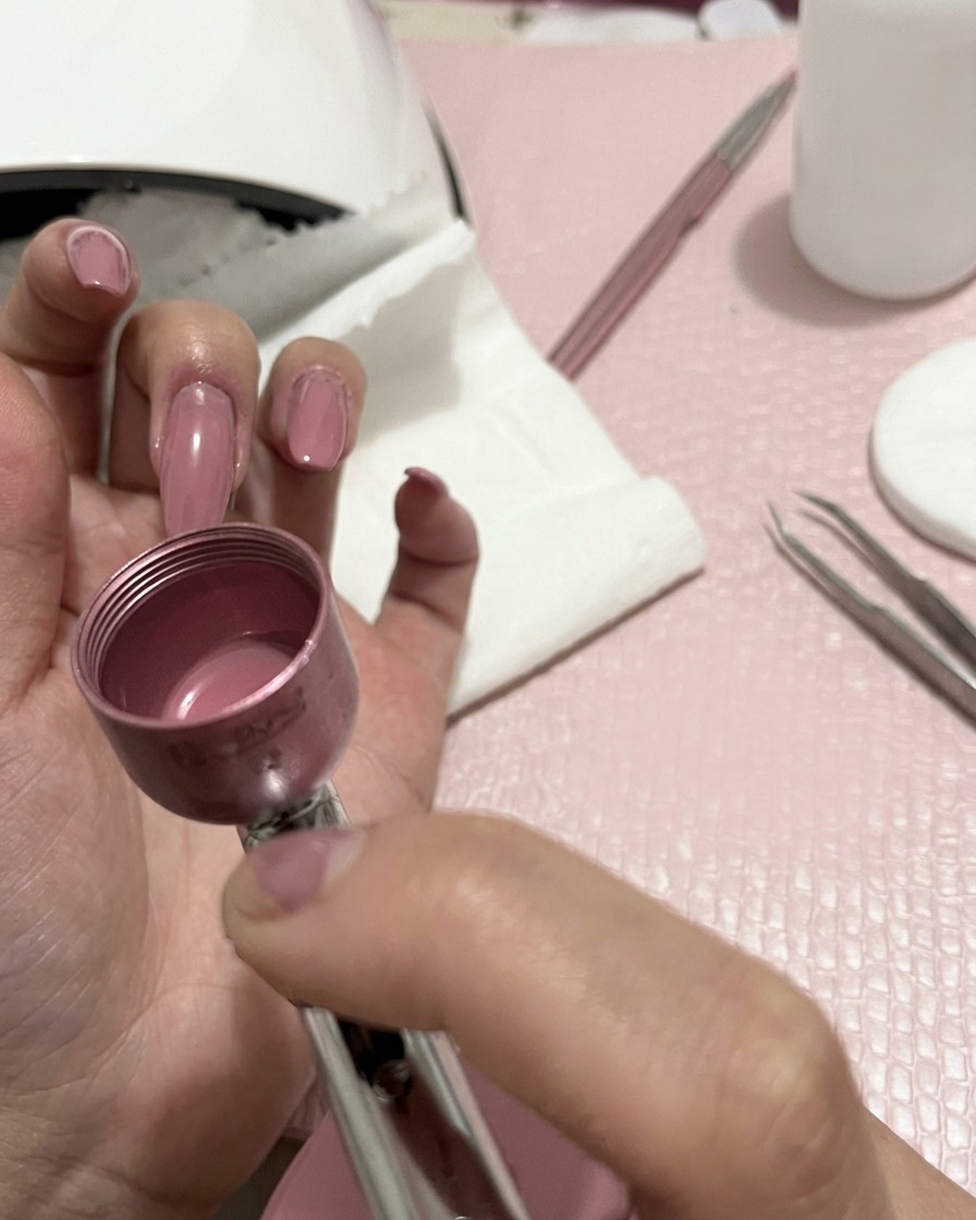A close-up of a hand holding an airbrush device, spraying a fine peach-colored mist onto manicured fingernails.