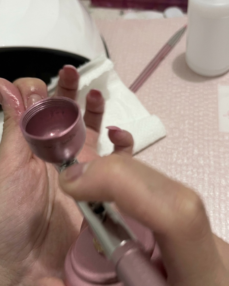 A close-up view of hands holding an airbrush, with the nozzle focused on blending the peach color on the nails.
