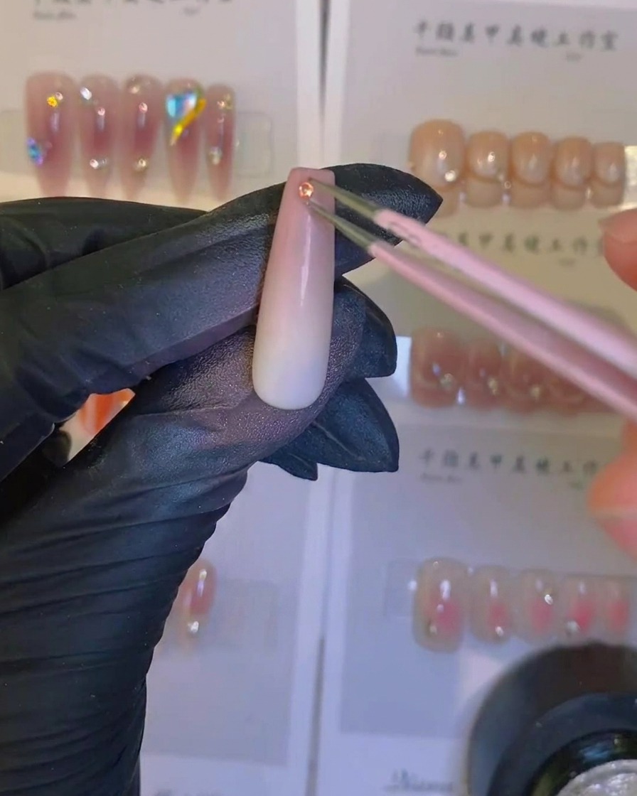 A brush applying a clear, glossy top coat over an airbrushed ombre nail with a crystal embellishment.