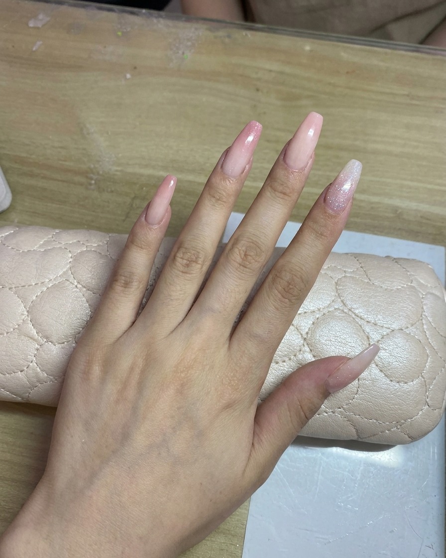 A hand with long, light-colored nails being airbrushed with a delicate pink gradient.