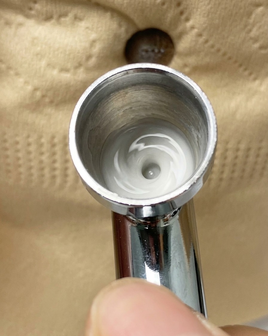 An airbrush spraying a fine mist of white pigment onto a nail tip that is partially covered with adhesive tape.